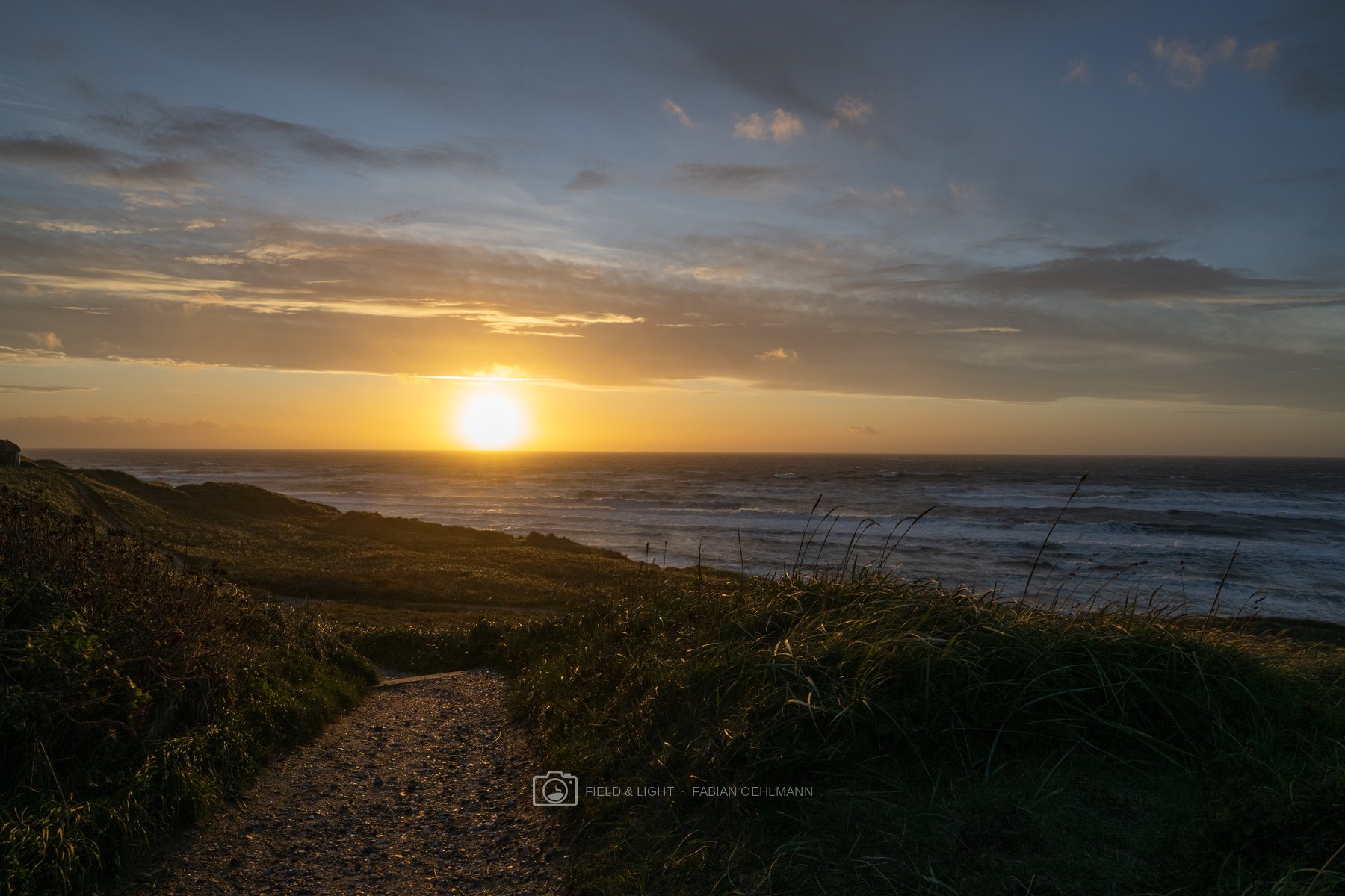 A narrow gravel path winding through dune grass towards the setting sun above the North Sea